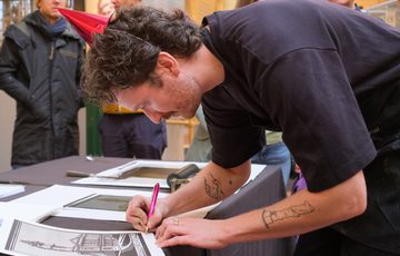 Person signs a print artwork at a table, while others observe.