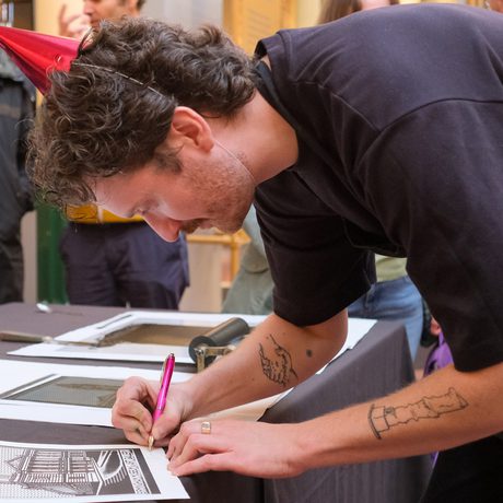 Person signs a print artwork at a table, while others observe.