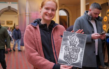 Smiling person holds a black-and-white print in a busy indoor setting.
