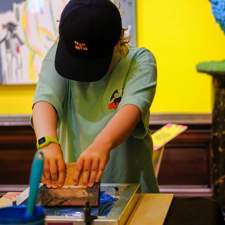 Child wearing a cap concentrates on screen printing with colourful art around.