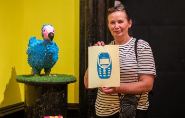 Woman holding artwork of a blue mobile phone, standing beside a colourful blue Dodo bird sculpture.