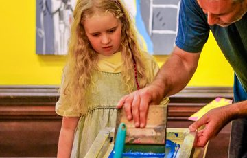 A man teaches a young girl screen printing with colourful abstract art in background.