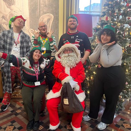 A group of staff dressed in festive jumpers and hats are next to Santa in a cafe.
