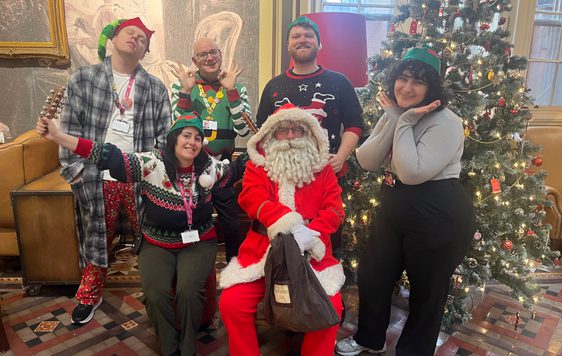 A group of staff dressed in festive jumpers and hats are next to Santa in a cafe.