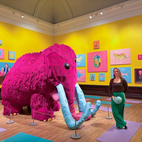 A person holding a pilates ball is standing in a bright colourful art gallery next to a large pink woolly mammoth.