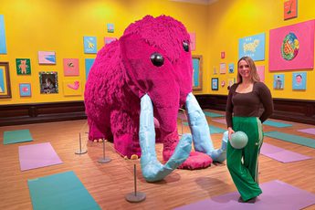 A person holding a pilates ball is standing in a bright colourful art gallery next to a large pink woolly mammoth.