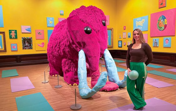 A person holding a pilates ball is standing in a bright colourful art gallery next to a large pink woolly mammoth.