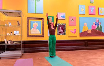 A person doing a standing pilates pose in a brightly lit gallery with colourful artwork on the walls.