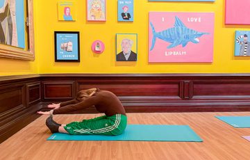 A person on the floor bending to touch their feet, doing pilates in a brightly lit gallery with colourful artwork on the walls.