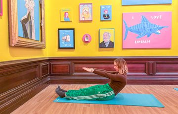 A person lying on the floor in a half sitting pose, doing pilates in a brightly lit gallery with colourful artwork on the walls.