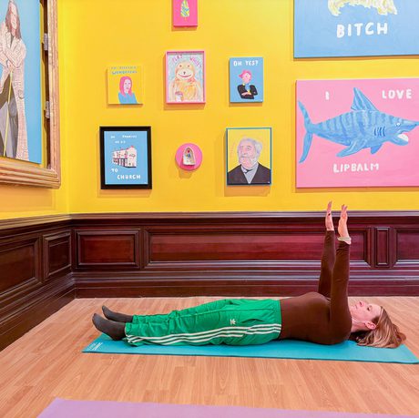 A person lying on the floor doing pilates in a brightly lit gallery with colourful artwork on the walls.