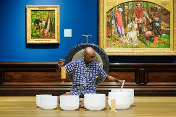 Man playing white crystal singing bowls in art gallery with paintings and a gong behind.
