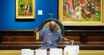 Man playing white crystal singing bowls in art gallery with paintings and a gong behind.