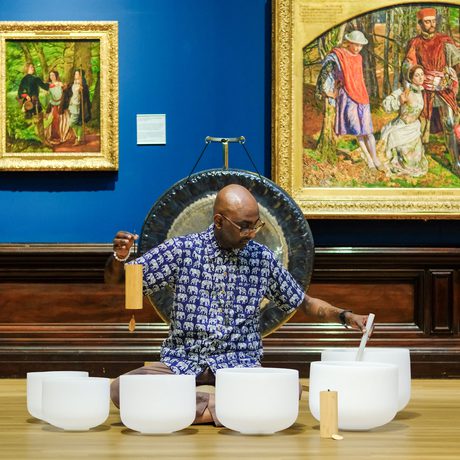 Man playing white crystal singing bowls in art gallery with paintings and a gong behind.