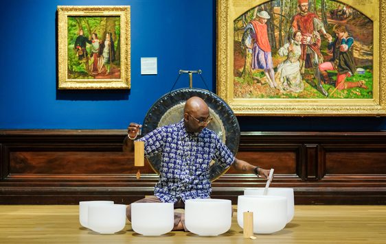 Man playing white crystal singing bowls in art gallery with paintings and a gong behind.