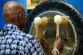 Man wearing elephant-patterned shirt plays large gong with two mallets against blue wall.