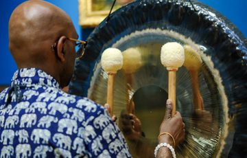 Man wearing elephant-patterned shirt plays large gong with two mallets against blue wall.
