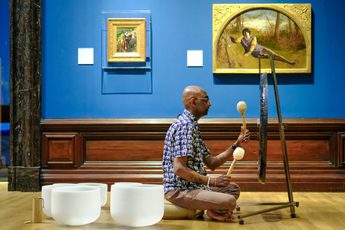 Man playing a gong with mallets, seated on the wooden floor in an art gallery.