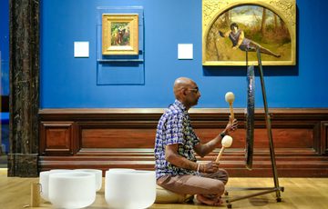 Man playing a gong with mallets, seated on the wooden floor in an art gallery.