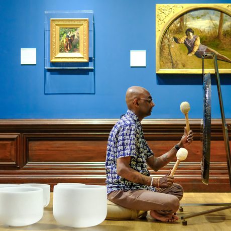 Man playing a gong with mallets, seated on the wooden floor in an art gallery.