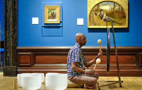 Man playing a gong with mallets, seated on the wooden floor in an art gallery.