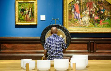Man plays gong and crystal bowls in art gallery with classical paintings on blue wall.