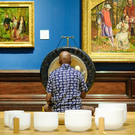 Man plays gong and crystal bowls in art gallery with classical paintings on blue wall.
