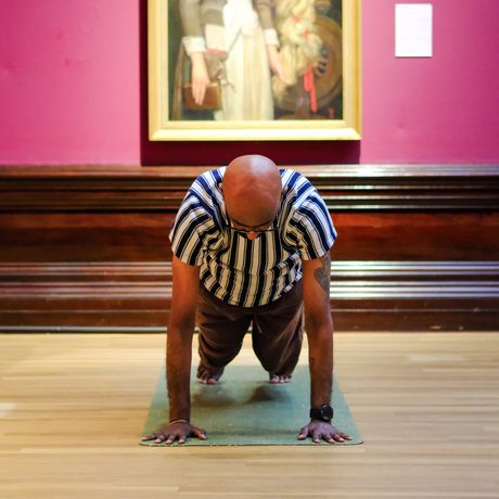 Man in striped shirt doing yoga on mat, with classical painting backdrop.