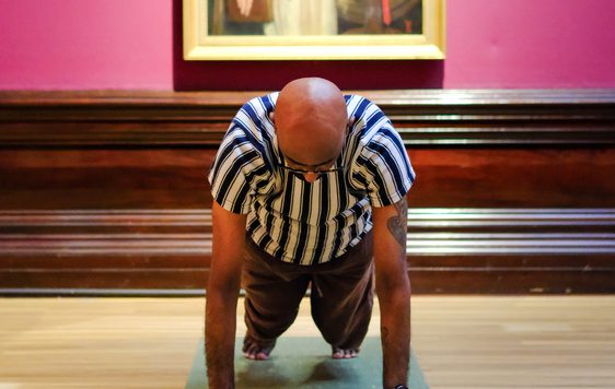 Man in striped shirt doing yoga on mat, with classical painting backdrop.