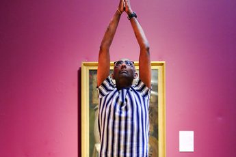Person in striped shirt performs yoga pose on mat against pink museum wall.