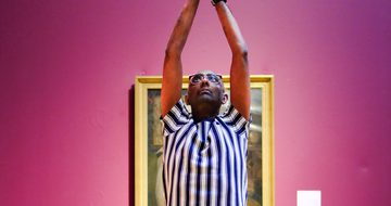 Person in striped shirt performs yoga pose on mat against pink museum wall.
