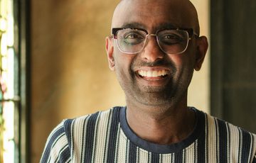 Smiling bald man with glasses wearing a striped shirt in softly lit indoor setting.