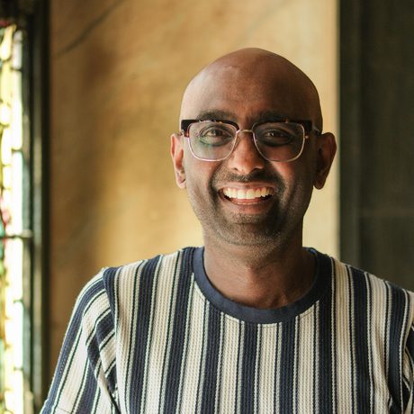 Smiling bald man with glasses wearing a striped shirt in softly lit indoor setting.