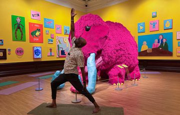 Man strikes a yoga pose in front of a large pink mammoth in an art gallery.