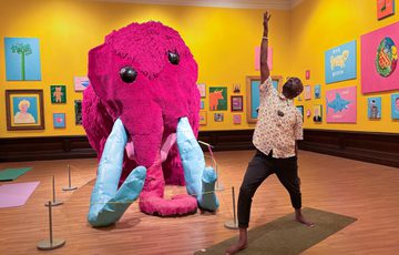Man strikes a yoga pose in front of a large pink mammoth in an art gallery.
