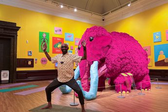 Man strikes a yoga pose in front of a large pink mammoth in an art gallery.