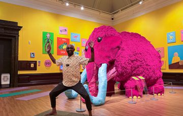 Man strikes a yoga pose in front of a large pink mammoth in an art gallery.