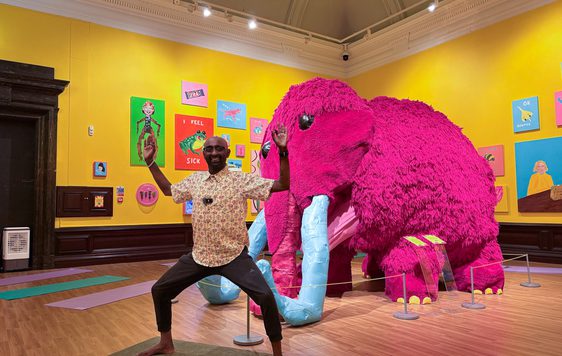 Man strikes a yoga pose in front of a large pink mammoth in an art gallery.