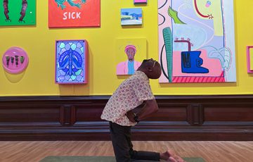 Man strikes a yoga pose in a colourful art gallery with paintings on a yellow wall.