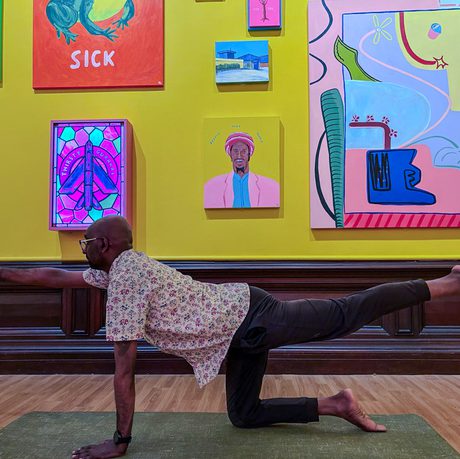 Man strikes a yoga pose in a colourful art gallery with paintings on a yellow wall.