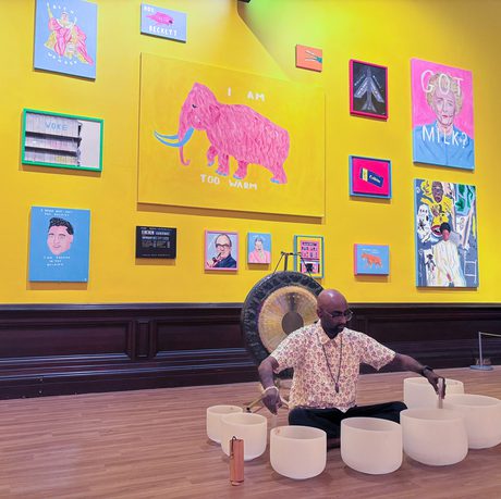 Man plays crystal bowls in a vibrant colourful art gallery with paintings on a yellow wall in background.