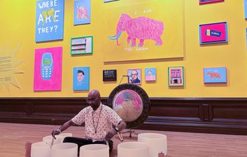 Man plays crystal bowls in a vibrant colourful art gallery with paintings on a yellow wall in background.
