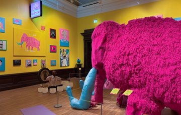 Man plays gong and crystal bowls in art gallery with vibrant colourful paintings on a yellow wall and in front of a large pink woolly mammoth.