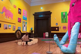 Man plays gong and crystal bowls in art gallery with vibrant colourful paintings on a yellow wall and in front of a large pink woolly mammoth.