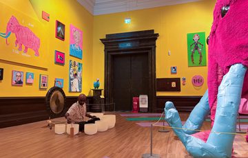Man plays gong and crystal bowls in art gallery with vibrant colourful paintings on a yellow wall and in front of a large pink woolly mammoth.