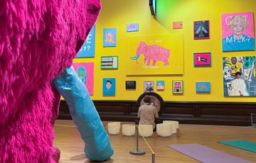 Man plays gong and crystal bowls in art gallery with vibrant colourful paintings on a yellow wall and part of pink woolly mammoth in shot.