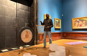 Man standing beside a gong and crystal singing bowls in an art gallery room.