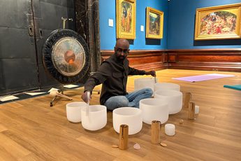 Man seated on wooden floor playing singing bowls with mallets, surrounded by Pre-Raphaelite paintings.