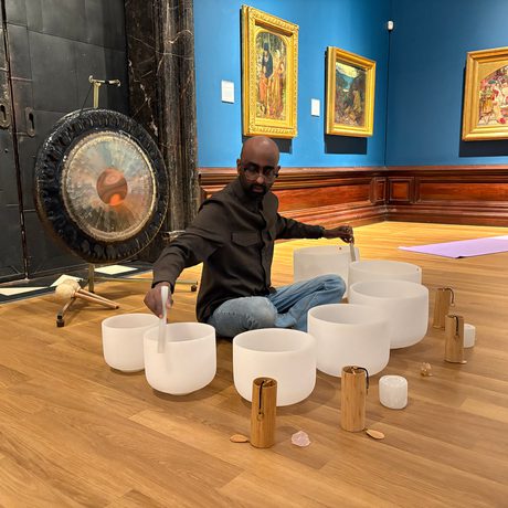 Man seated on wooden floor playing singing bowls with mallets, surrounded by Pre-Raphaelite paintings.