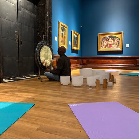 Person plays a gong in an art gallery, surrounded by singing bowls, yoga mats and Pre-Raphaelite paintings on the walls.